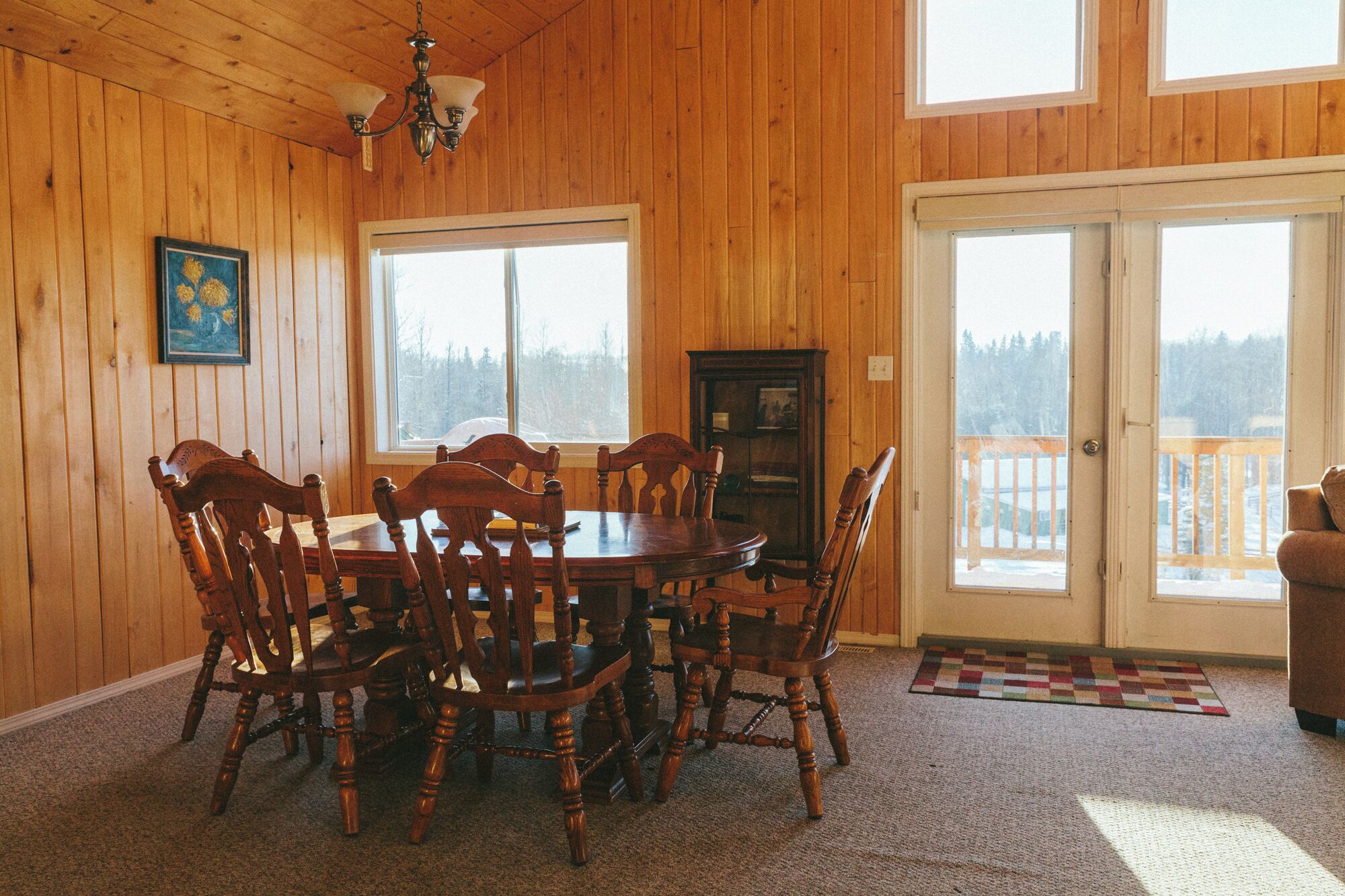 Ein Esszimmer mit Holzwandverkleidung, einem runden Tisch, sechs Stühlen, einer Chandelierleuchtre, einem Schrank und einem großen Fenster mit Blick auf eine verschneite Landschaft.