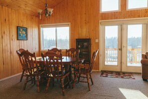 A wooden-paneled dining room with a round table and six chairs, a chandelier, a cabinet, and a large window with a view of a snowy landscape.