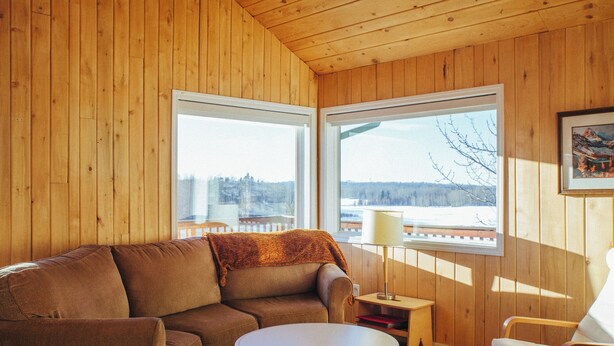 A cozy room with a brown leather sofa, a small round table, and a wooden chair. There are two windows providing a view of a snowy landscape.