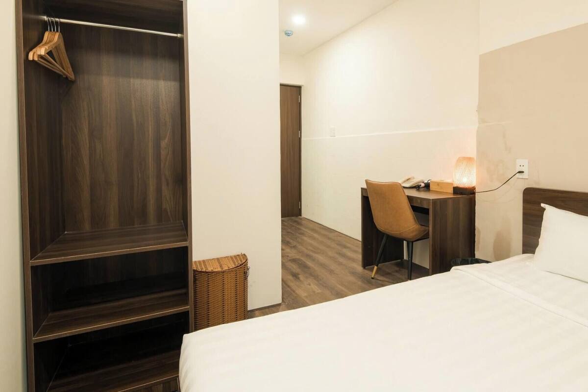 A hotel room with a bed, a desk with a chair, a wardrobe, and a bedside lamp.