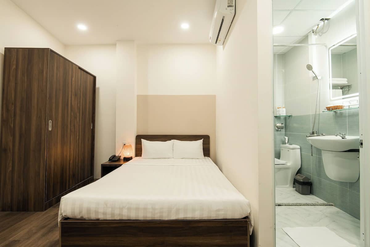 A hotel room with a bed, wardrobe, bedside table, and a bathroom with a sink, toilet, and mirror.