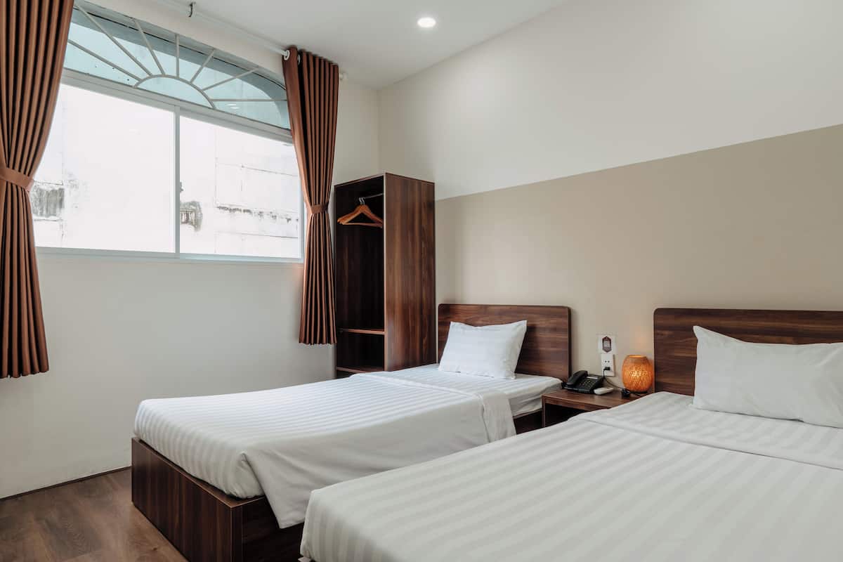A hotel room with two beds, a wooden wardrobe, a bedside table with a lamp, and a window with curtains.