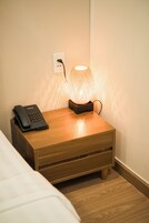 A wooden bedside table with a lamp, a phone, and a power outlet on the wall.