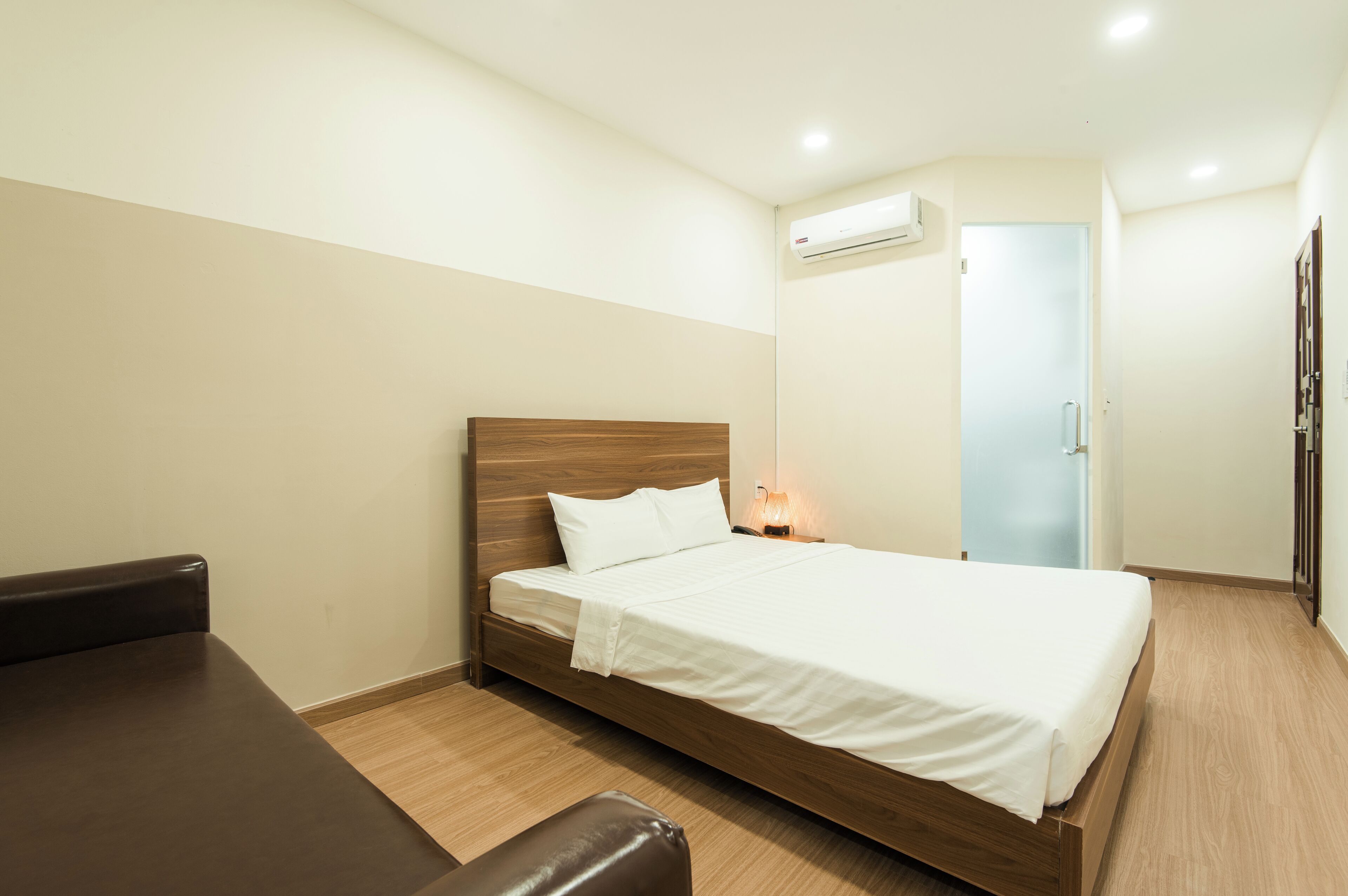 A hotel room with a wooden bed, a bedside table, a chair, and an air conditioning unit on the wall.