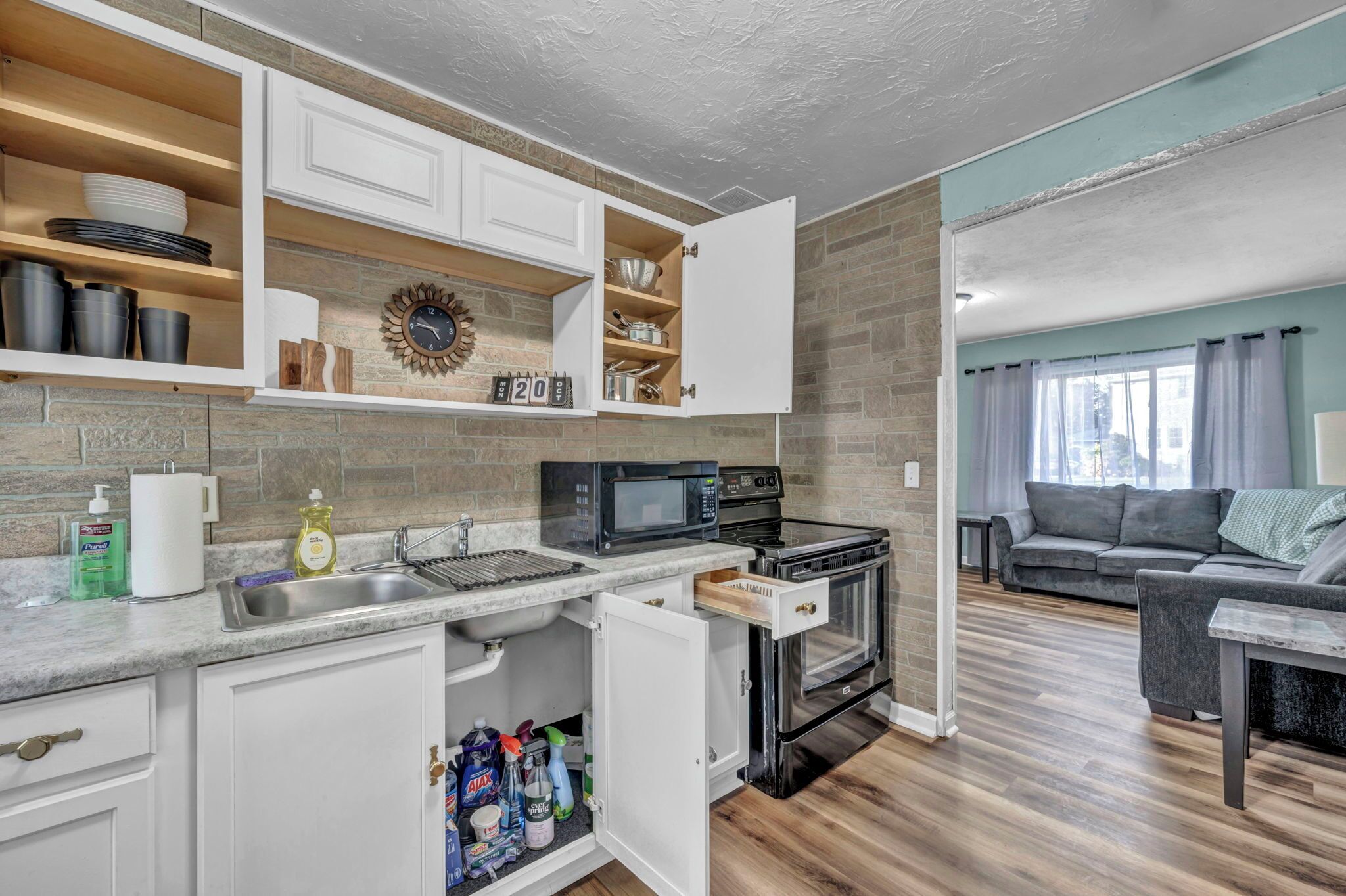 A compact kitchen with white cabinets, a microwave, and an oven. There is a dining area with a gray sofa and a small table.