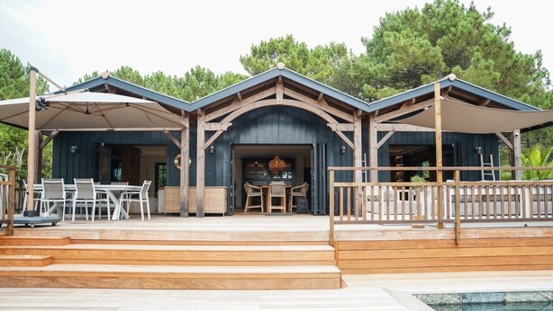 Un área de piscina moderna al aire libre con escalones de madera que llevan a un porche cubierto con mobiliario exterior.
