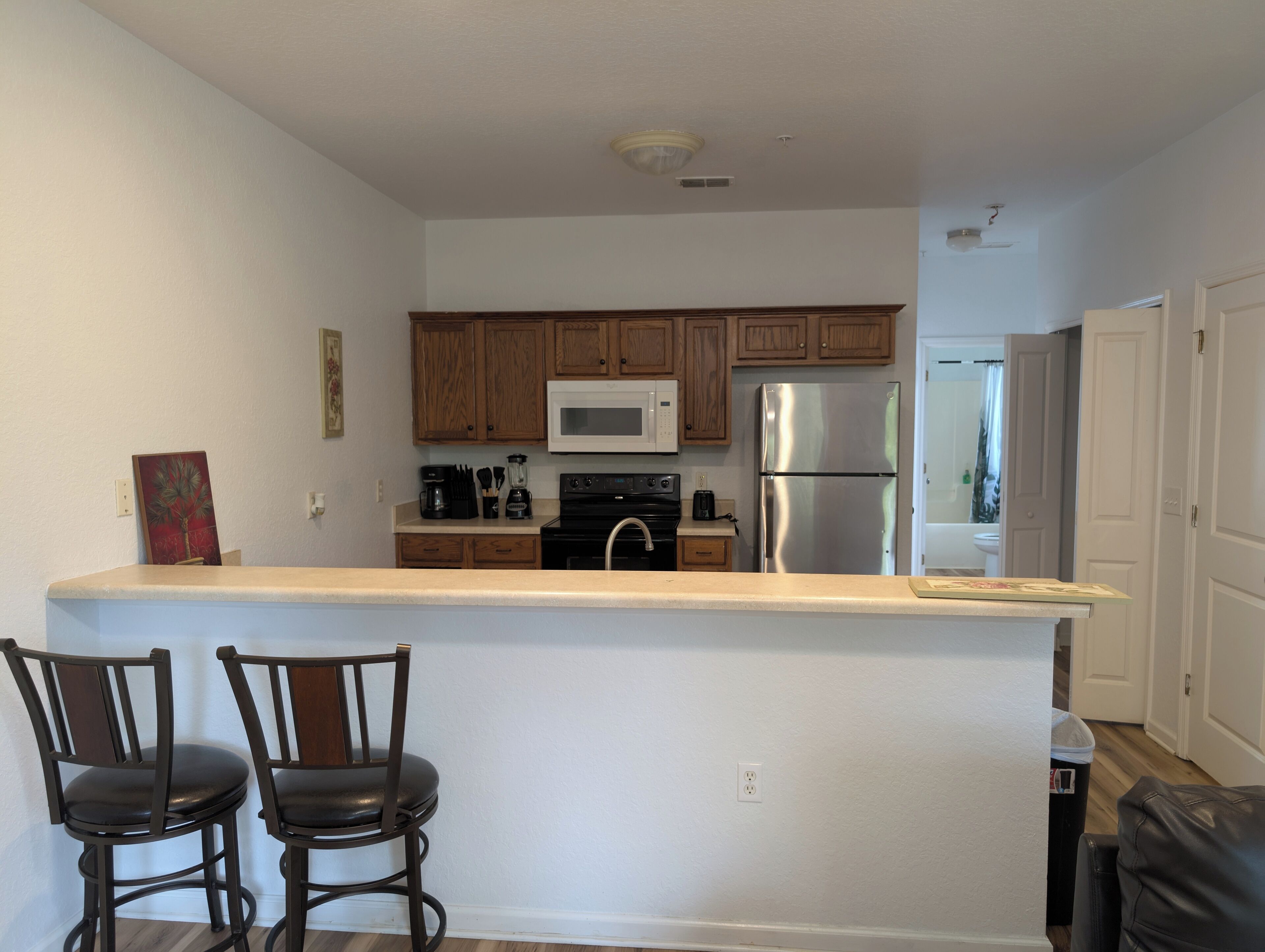 A kitchen with wooden cabinets, a stainless steel refrigerator, a microwave, and a coffee maker. There are two chairs at the counter.