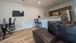 A modern living room with a leather sofa, a dining table with chairs, a ceiling fan, a flat-screen TV mounted on the wall, and a kitchen area with wooden cabinets and stainless steel appliances.