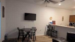 A modern living room with a dining area, a ceiling fan, a flat-screen TV, and a kitchen counter.