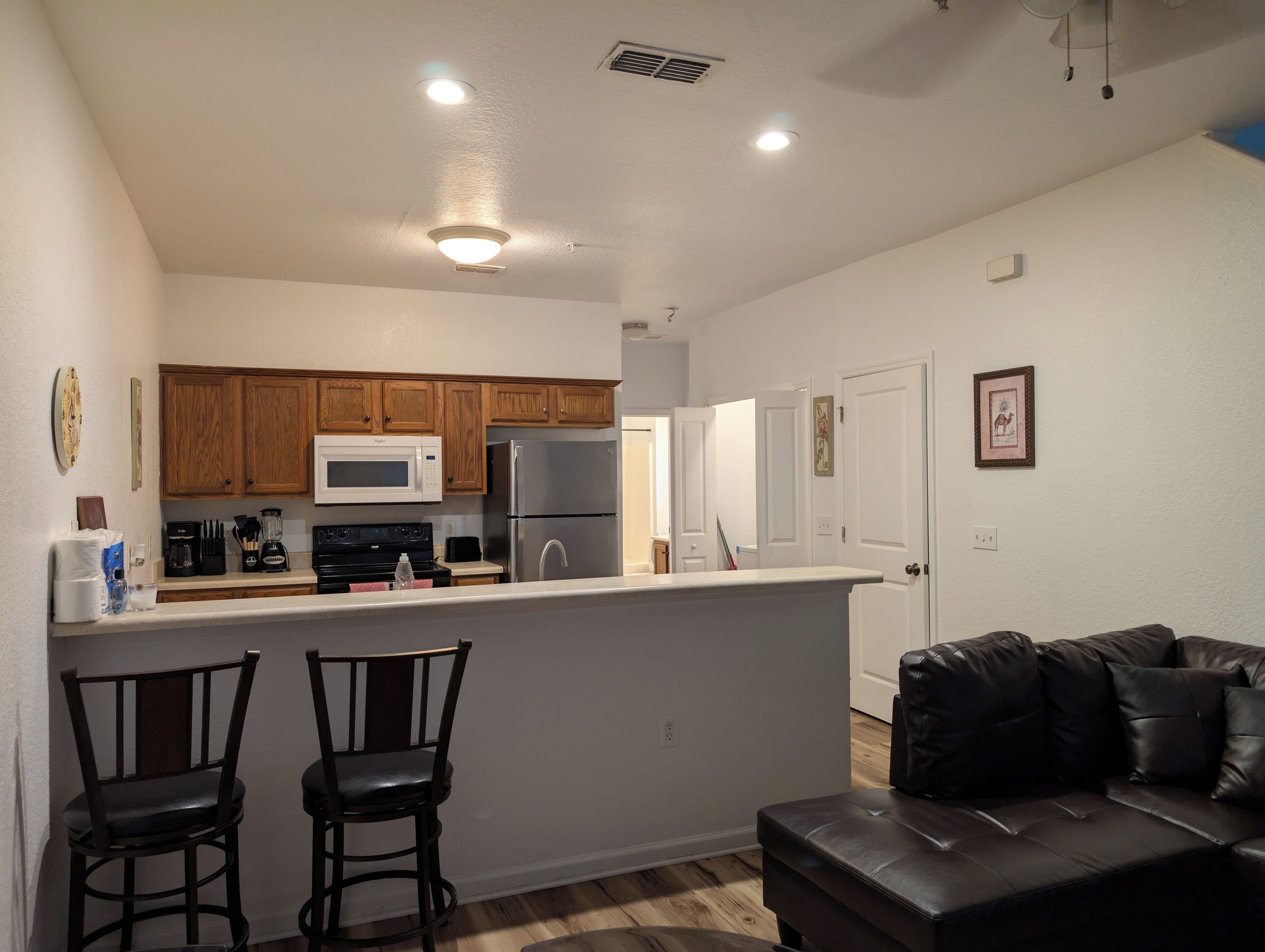 A compact kitchen with wooden cabinets, a microwave, a refrigerator, and a bar area. There are two bar stools and a black leather couch in the living area.