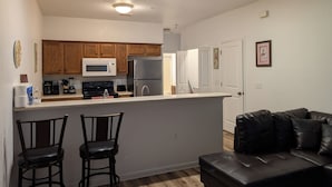 A compact kitchen with wooden cabinets, a microwave, a refrigerator, and a bar area. There are two bar stools and a black leather couch in the living area.