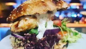 A burger with sesame seed bun, lettuce, red onion, and a creamy sauce, served on a white plate. - Gasthaus Sofram (Frastanz)