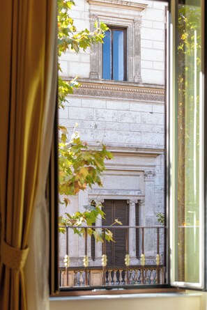 Ein Fenster mit Blick auf eine Gebäudefassade, inklusive Balkon mit Geländer und einem Fenster mit Rollläden.