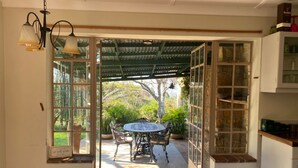 An open doorway leading to an outdoor patio with a round table and chairs, surrounded by potted plants and a view of trees. - Victorian style cottage in Nyanga - 2096 (Nyanga)