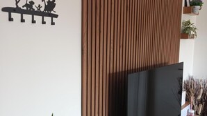 A modern living room with a flat-screen TV on a wooden console, a wall-mounted shelf with decorative items, and a large green plant. - Hibiscus Apartment (Ciampino/Rome/Roma) (Ciampino)