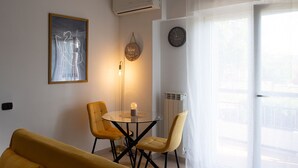 A modern living room with a round glass table, two yellow chairs, a framed abstract artwork, a hanging light fixture, a wall clock, and a white radiator. - Hibiscus Apartment (Ciampino/Rome/Roma) (Ciampino)