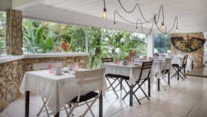 Ein Restaurant mit weißen Tischen und Stühlen, Steinwänden und großen Fenstern mit Grünpflanzen davor.