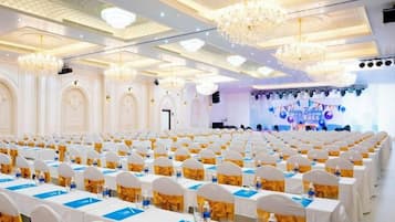 Un salón de banquetes con filas de mesas, candelabros y un escenario con fondo azul.