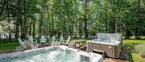 An outdoor hot tub area with Adirondack chairs and a picnic table, surrounded by a forest.