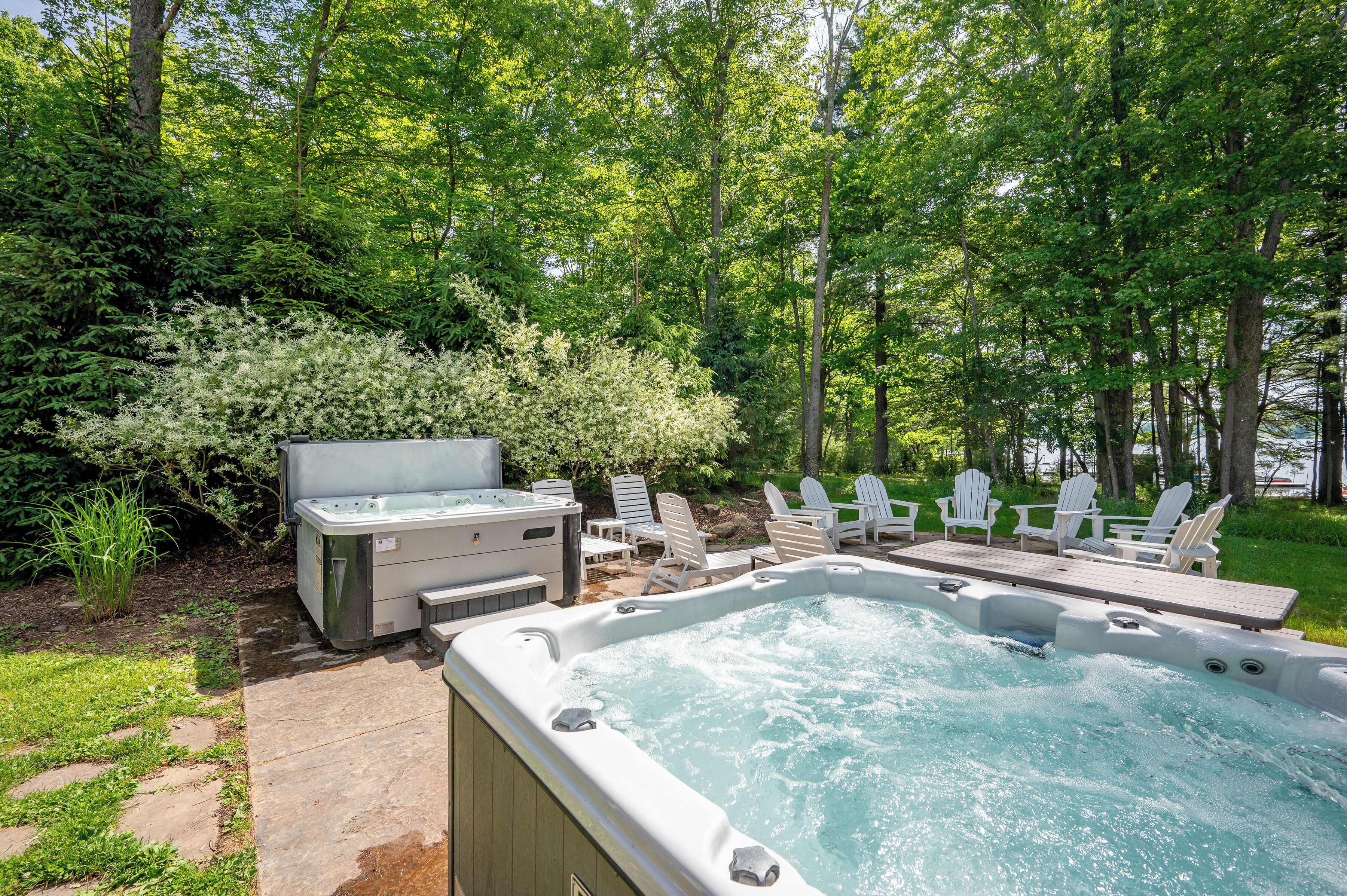 A hot tub with a wooden deck and white Adirondack chairs, surrounded by lush greenery and trees.