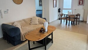 A living room with a sofa, a coffee table, a dining area with a table and chairs, a clock on the wall, and a window.
