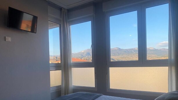 A bedroom with a bed, a TV mounted on the wall, and large windows offering a mountain view. - Pension San Vicente (San Vicente de la Sonsierra)