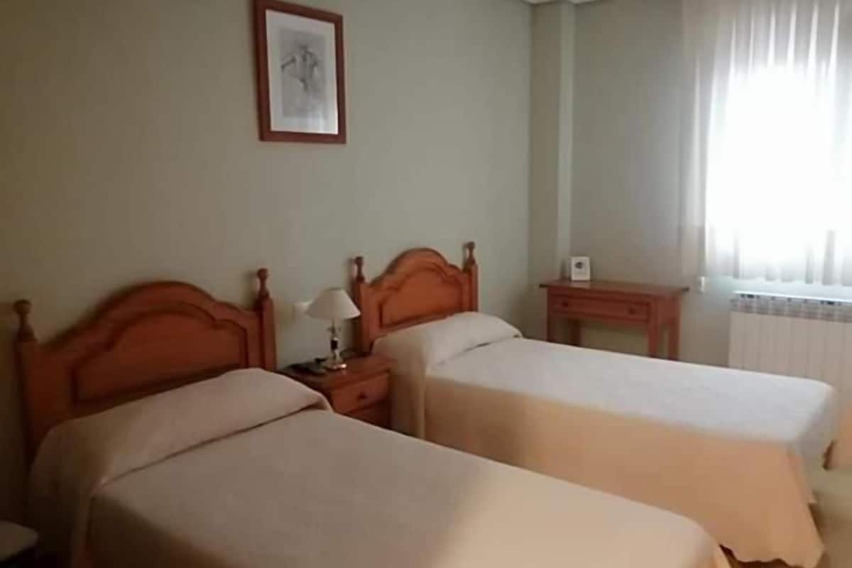 Ein Hotelzimmer mit zwei Betten, einem Nachttisch mit Lampe, einem gerahmten Bild an der Wand und einem Fenster mit Vorhängen.