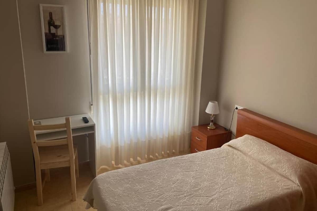 Ein Schlafzimmer mit einem Bett, einem Schreibtisch, einem Stuhl, einem Nachttisch und einem Fenster mit durchsichtigen Vorhängen.
