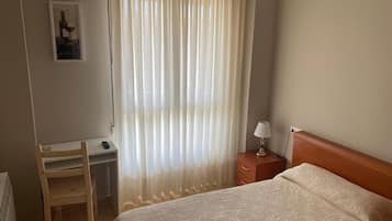 A bedroom with a bed, a desk, a chair, a nightstand, and a window with sheer curtains.
