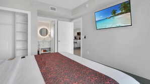 Quarto de hotel com uma cama, um guarda-roupa, casa de banho com espelho e uma televisão exibindo uma cena de praia.