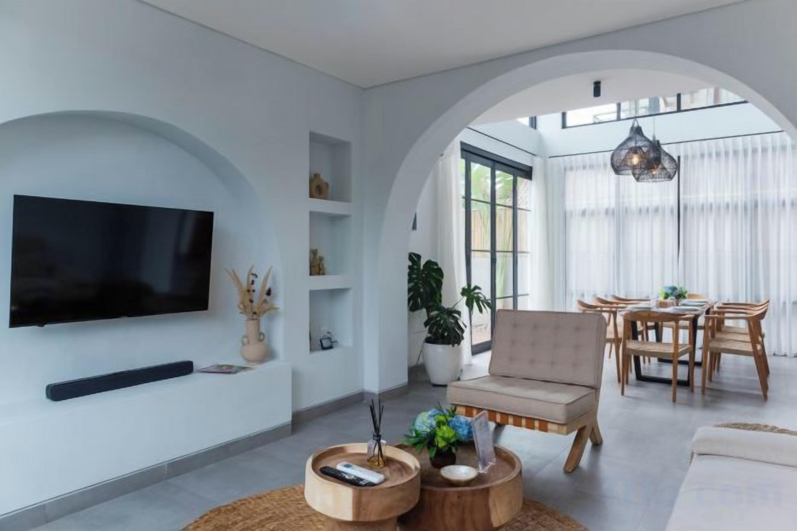 A modern living room with a flat-screen TV, a sofa, a dining area with a table and chairs, and a large arched window.