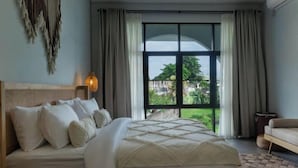 Een slaapkamer met een groot bed, hoofdeinde, een raam met gordijnen en uitzicht op groen.