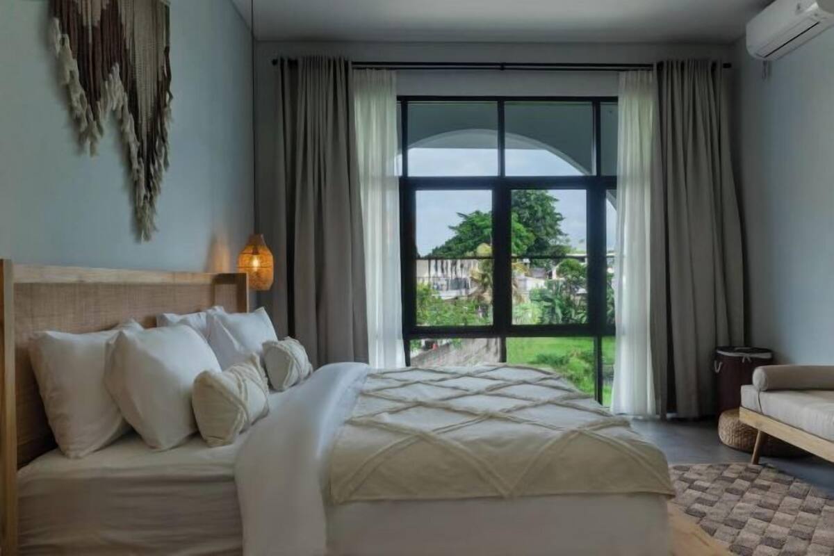 Een slaapkamer met een groot bed, hoofdeinde, een raam met gordijnen en uitzicht op groen.