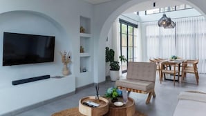Een moderne woonkamer met een flatscreentelevisie, een bank, een eethoek met houten stoelen en een groot raam met lichte gordijnen.