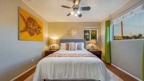 Ein Schlafzimmer mit einem Bett, zwei Nachttischen, einem Deckenventilator, einem Fenster mit Vorhängen und einem Bild an der Wand.