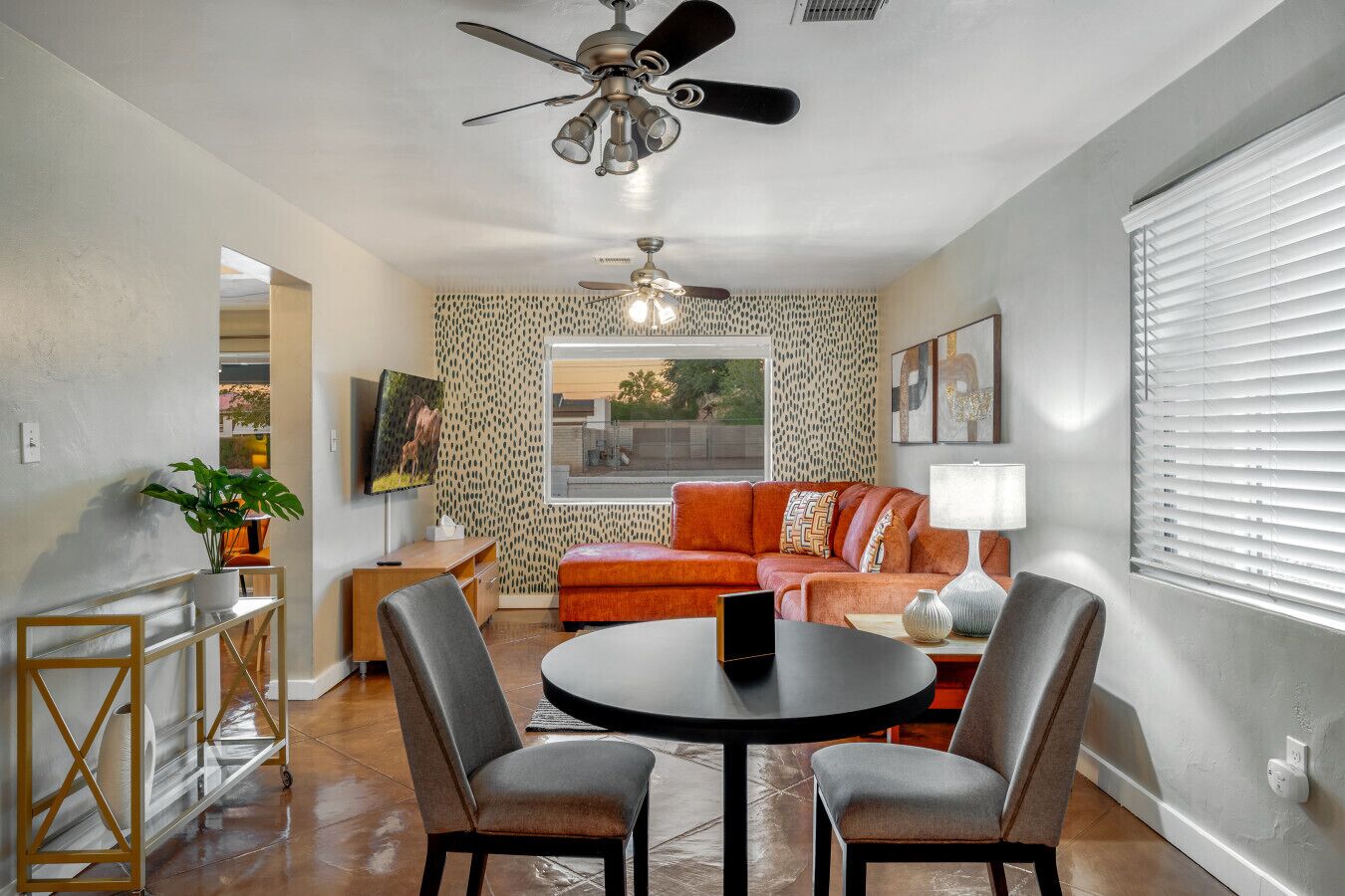 Uma sala de estar moderna com uma mesa redonda, duas cadeiras, um sofá, um ventilador de teto e uma janela grande com persianas.