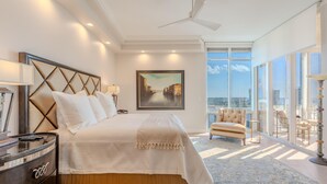 A spacious bedroom with a large bed, a nightstand, a chair, and a view of the city through large windows. - Palm Harbor #704 (Fort Myers Beach)