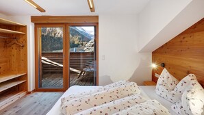 Une chambre avec une tête de lit en bois, un lit recouvert d’une literie à motifs floraux, un banc en bois et une porte-fenêtre coulissante donnant sur une terrasse extérieure.