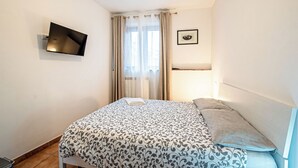 Ein Schlafzimmer mit einem Bett, einem flachbildfernseher an der Wand, einem Fenster mit Vorhängen und einem gerahmten Bild.