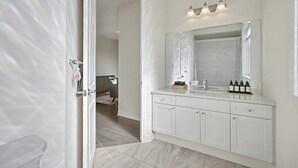 Un baño moderno con un espejo grande, muebles blancos y una zona de ducha visible a través de una puerta abierta.