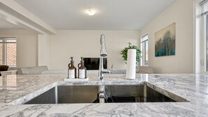 Una cocina moderna con una isla grande, un lavaplatos de acero inoxidable y un horno empotrado. Se observa un rincón de descanso con un sofá y una mesa de centro al fondo.