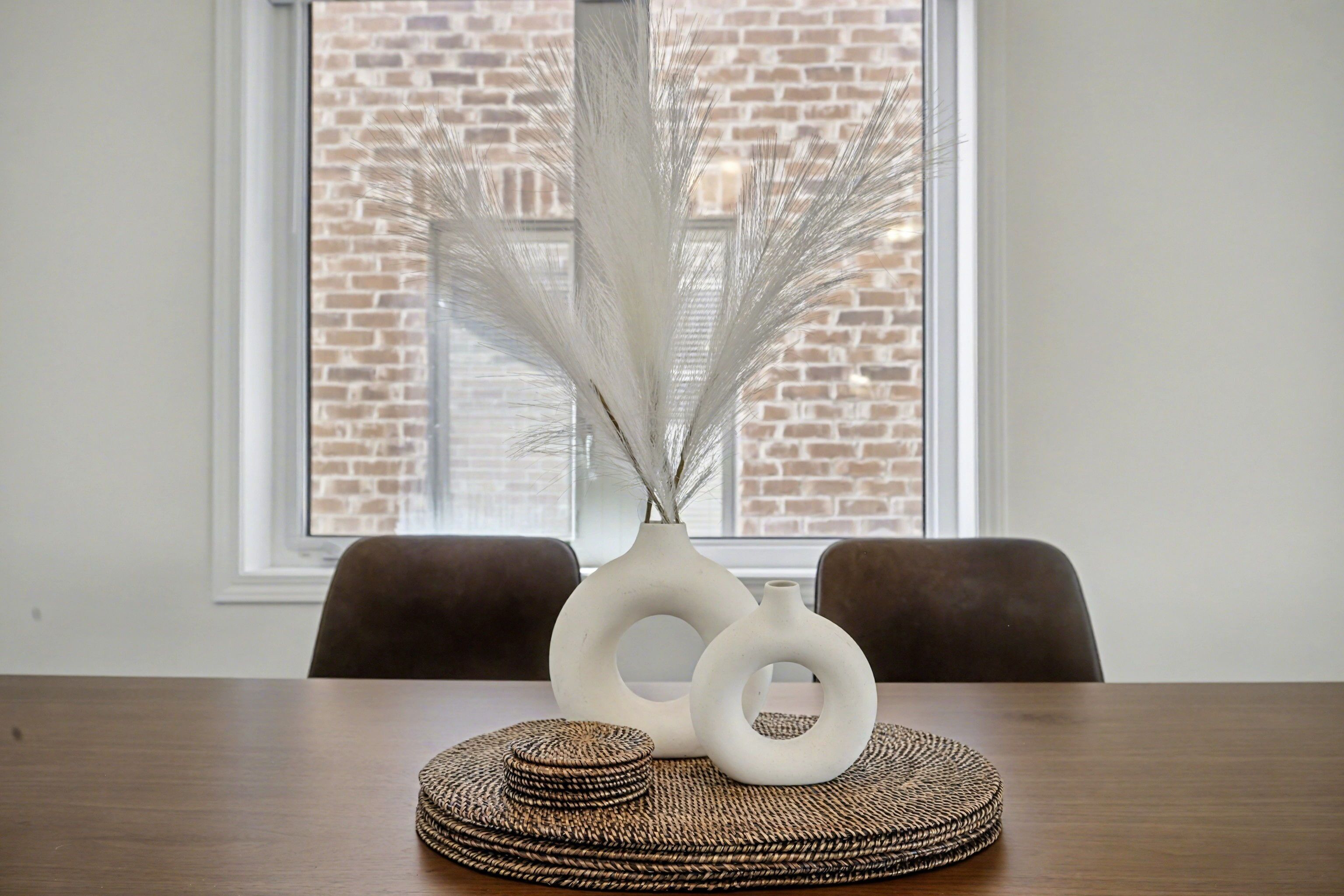 A dining table with a vase of white feathers, two white vases, and woven coasters.