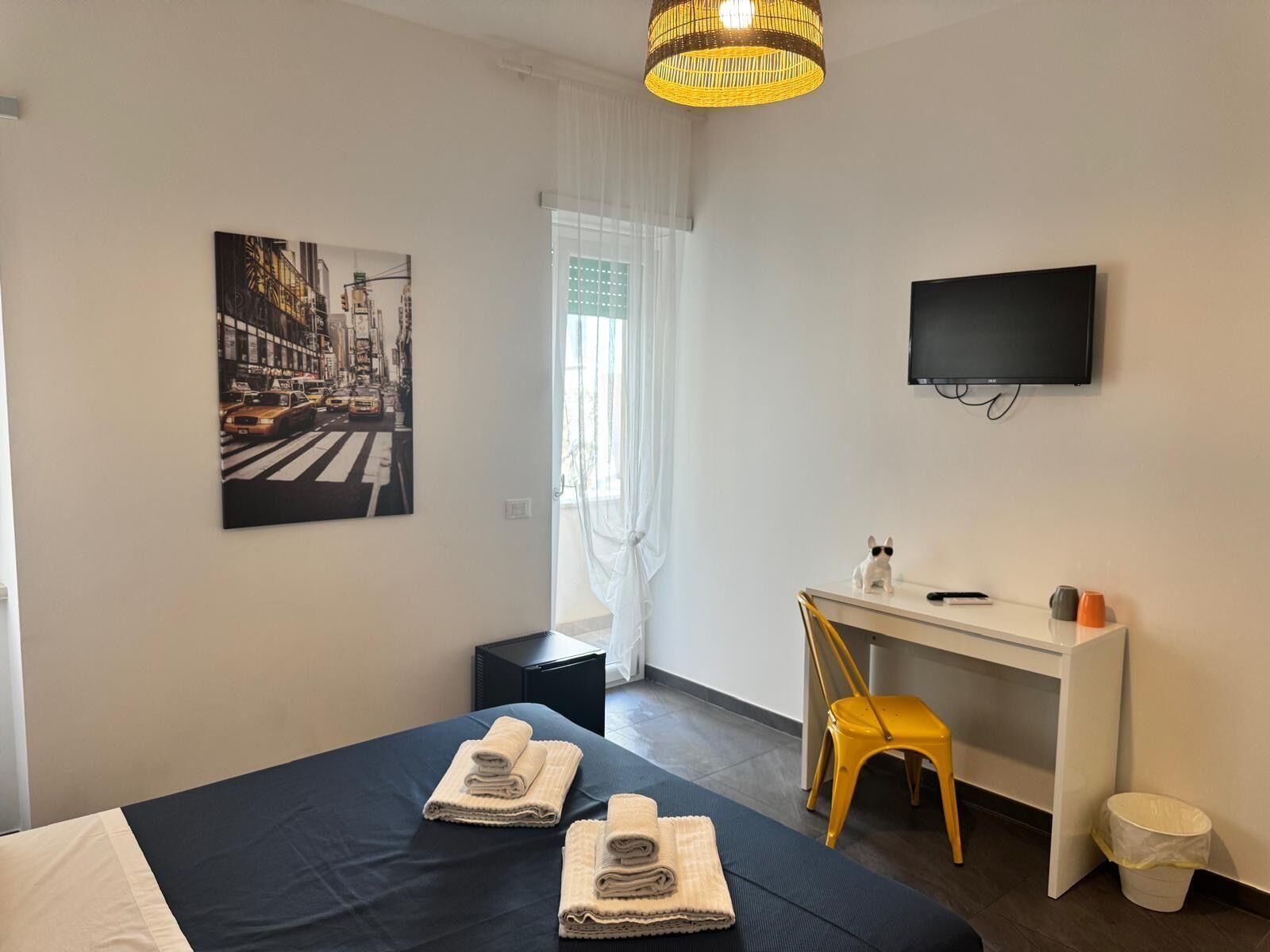 A hotel room with a bed, a desk, a chair, a TV, and a painting on the wall.