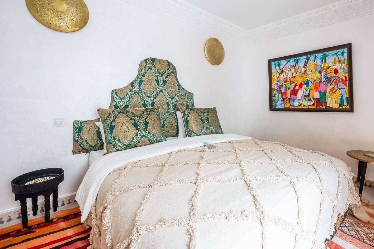 A bedroom with a bed, a painting, a small round table, and a decorative stool.