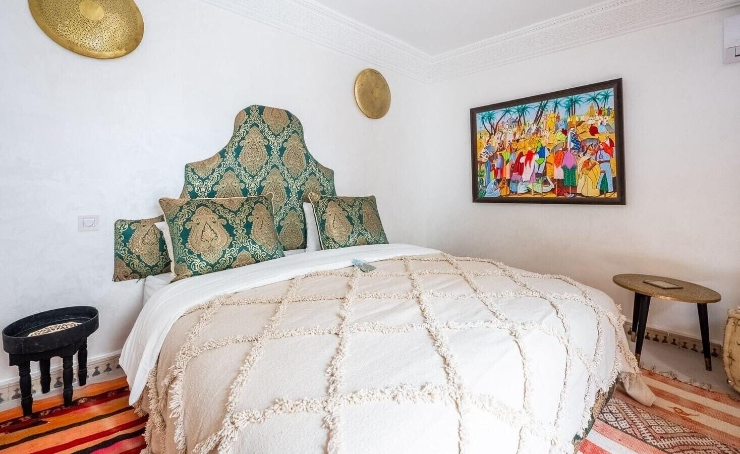 A bedroom with a large bed, patterned pillows, a painting on the wall, a small stool, and a decorative bowl.