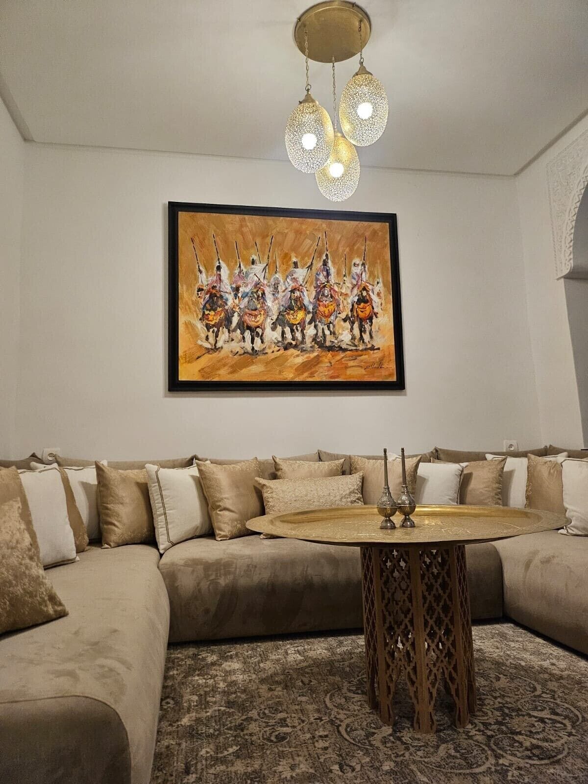 Un salon avec un canapé beige, une table basse ronde sur laquelle repose un plateau, et un tableau représentant un groupe de personnes à cheval.