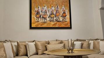 Uma sala de estar com um sofá bege, uma mesa de centro redonda com um tabuleiro e uma pintura de um grupo de pessoas a cavalo.
