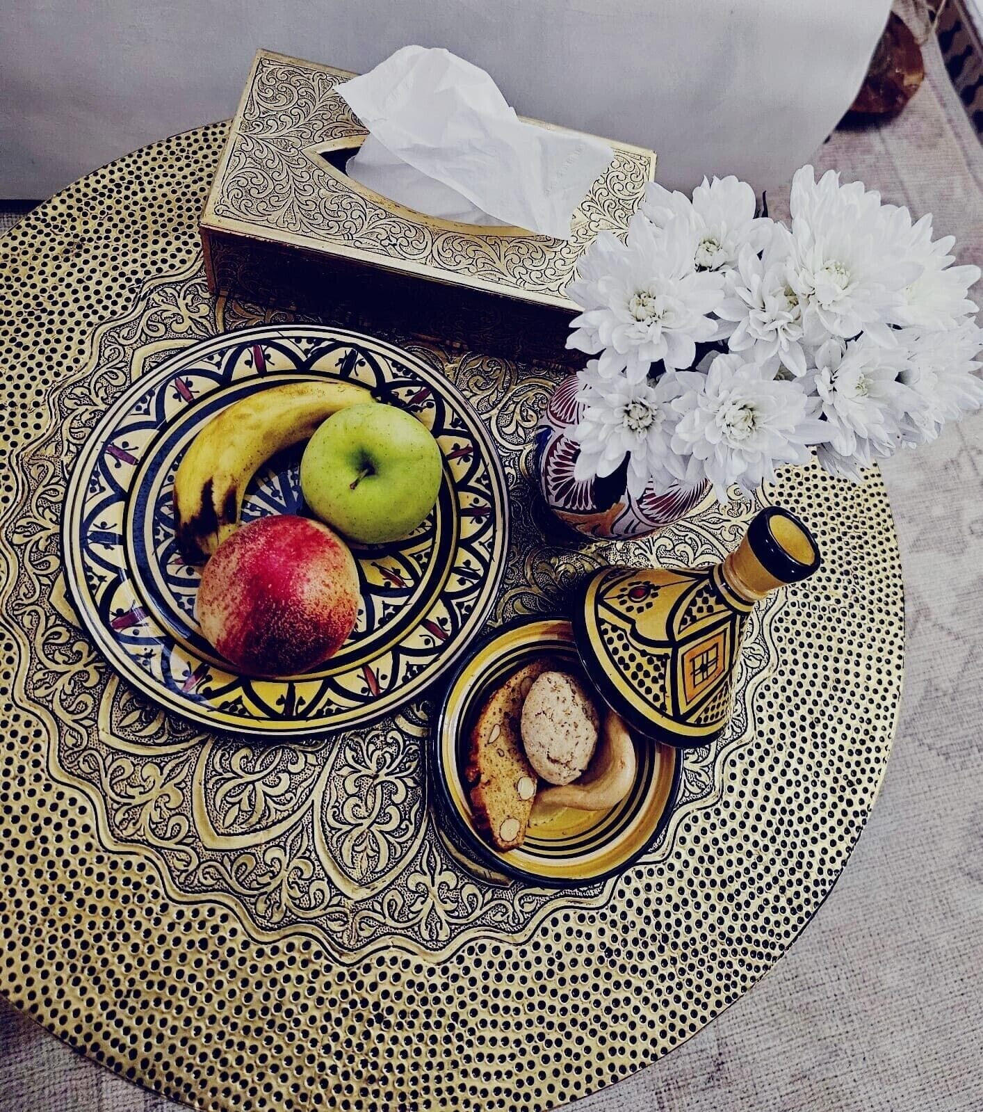 A tray with a plate of fruits, a small bowl of cookies, and a decorative bottle, all on a patterned mat.