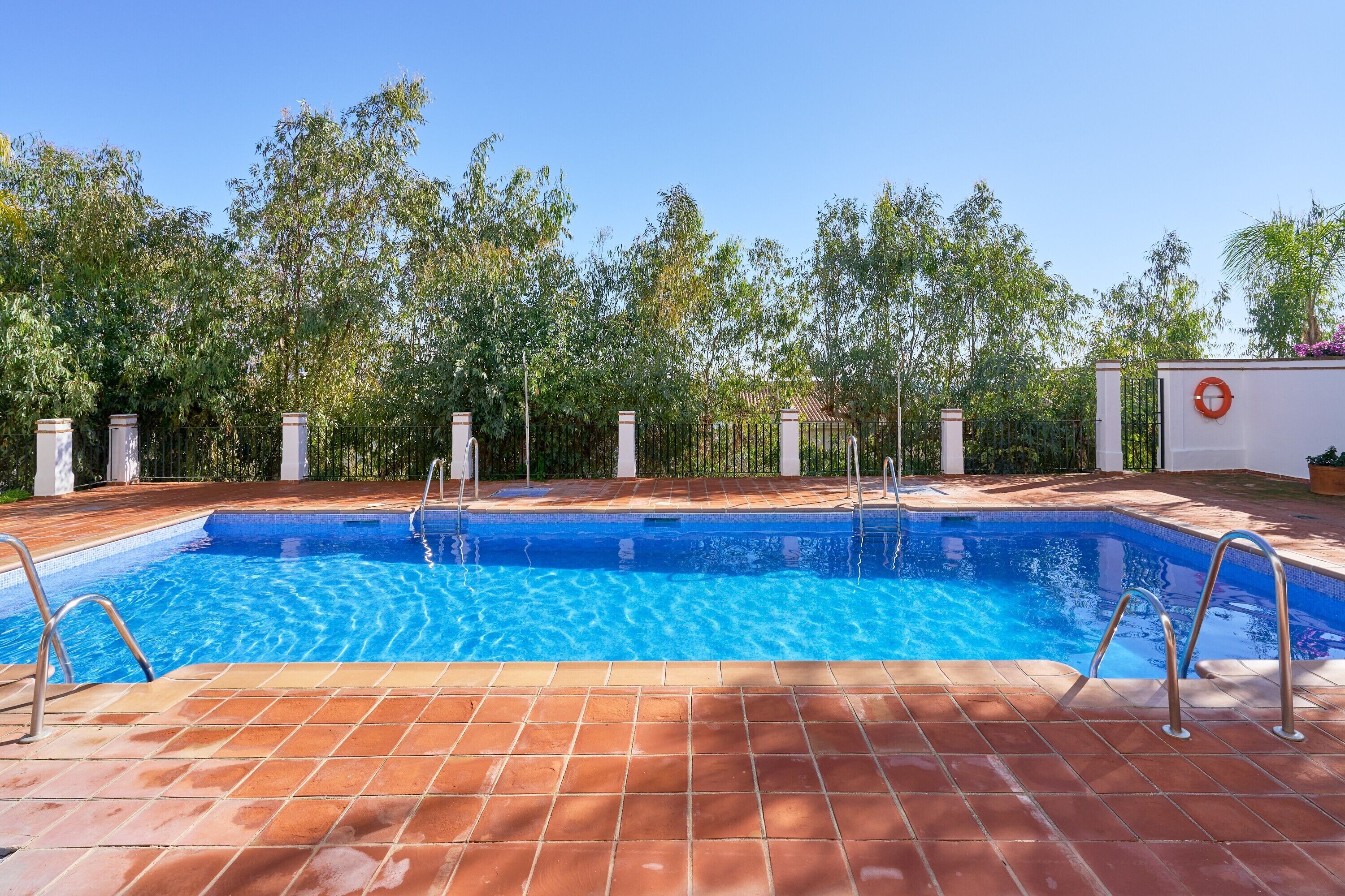 A rectangular swimming pool with clear blue water, surrounded by a tiled deck and a fence.