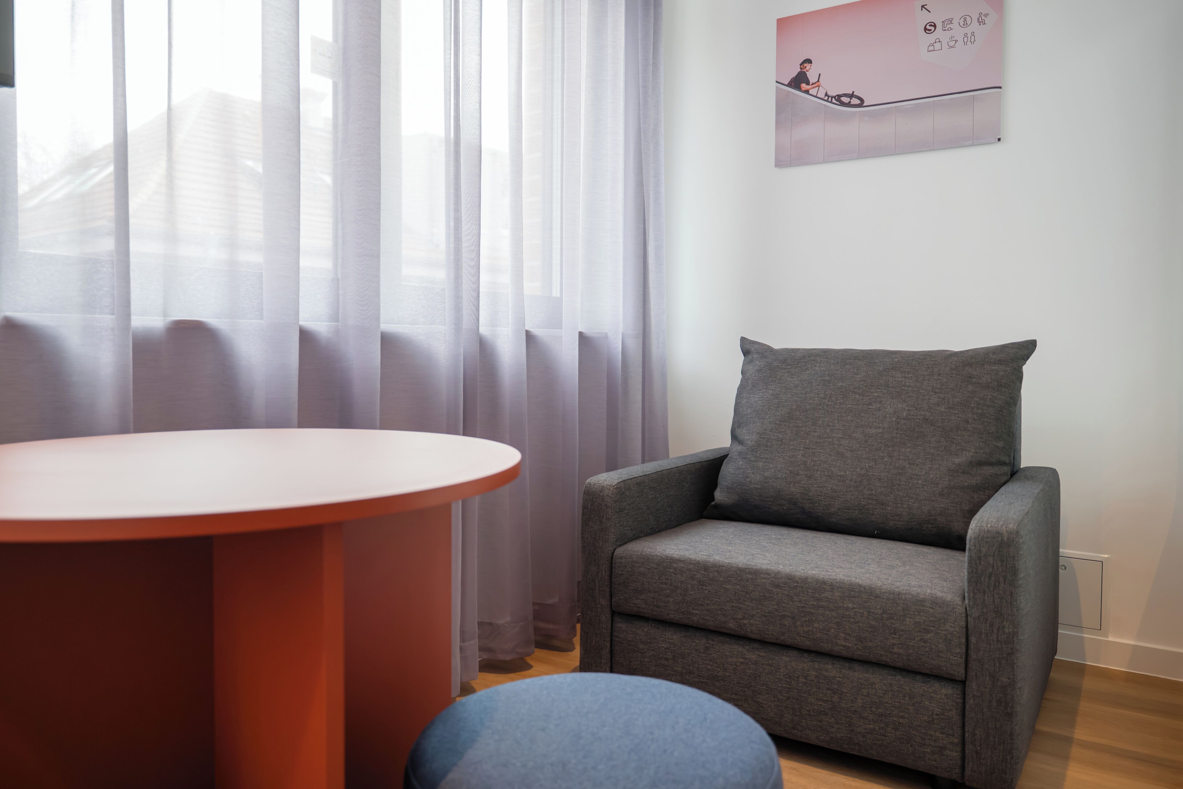 Habitació moderna amb una poltrona gris, una taula rodona i cortines transparents.
